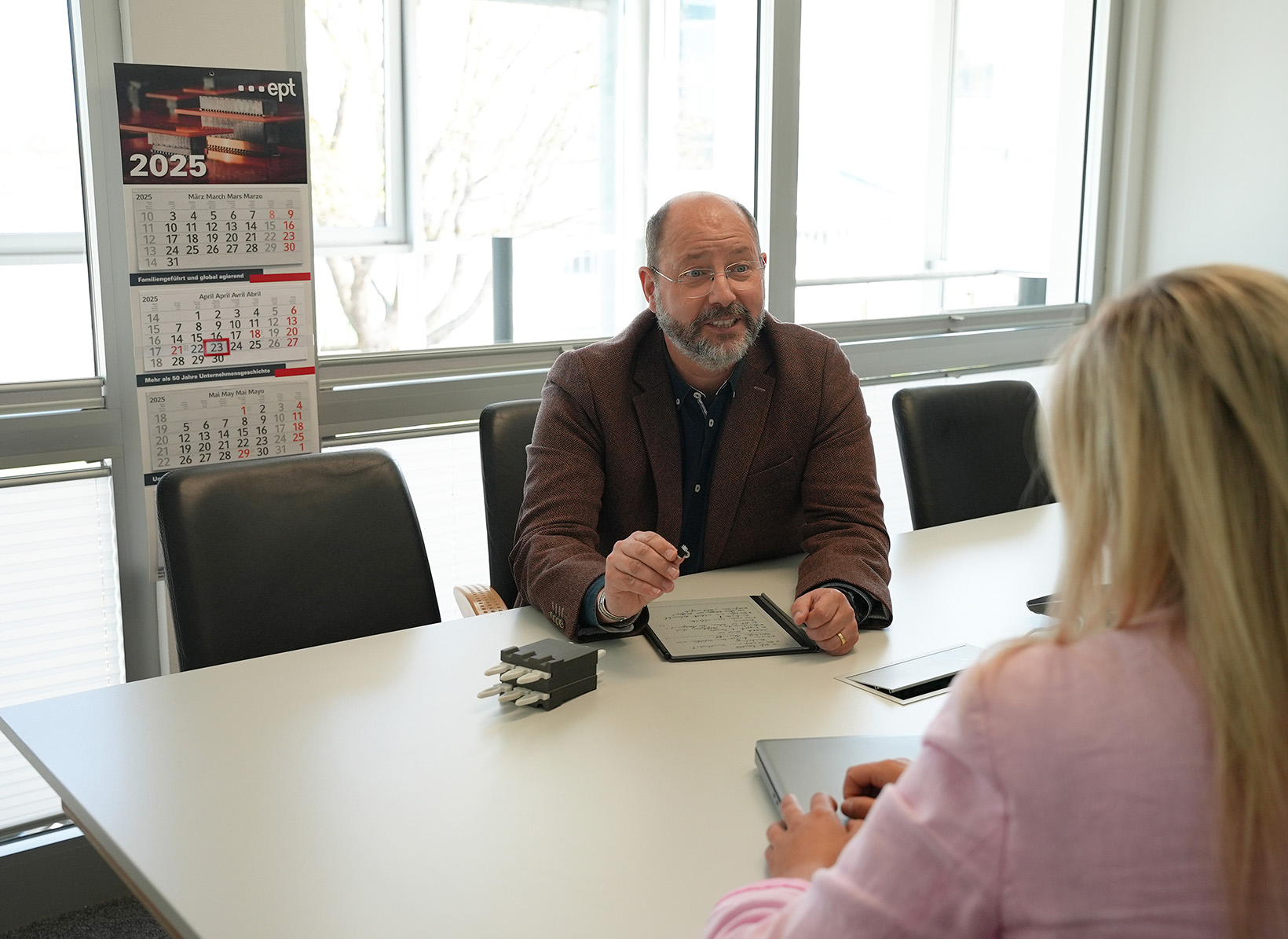 Auf dem Bild ist eine Besprechung zwischen zwei Personen in einem modernen Konferenzraum zu sehen, wobei ein Mann Notizen macht und die Frau ihm aufmerksam zuhört.