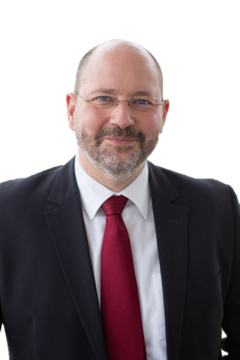 Auf dem Bild ist ein Mann mit Brille in einem Anzug und einer roten Krawatte zu sehen, der freundlich in die Kamera lächelt.