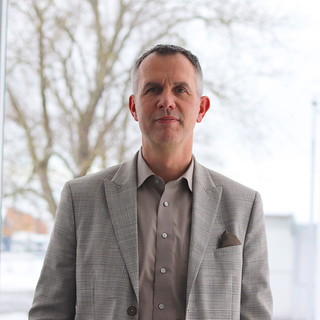 Auf dem Bild ist ein Mann in einem eleganten Anzug vor einem hellen, unscharfen Hintergrund zu sehen.