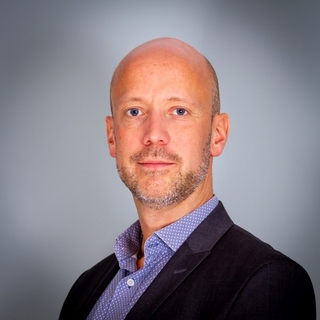 Auf dem Bild ist ein Mann mit Glatze und einem Bart zu sehen, der einen Anzug trägt und in die Kamera schaut.