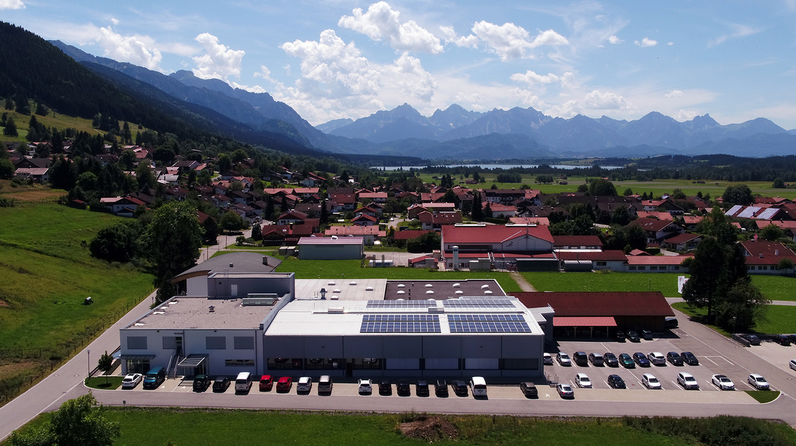 Auf dem Bild ist eine ländliche Szene zu sehen, die ein modernes Gebäude mit Photovoltaikanlagen sowie ein Wohngebiet vor einer Berglandschaft zeigt.