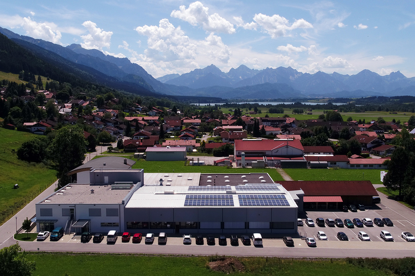 Auf dem Bild ist eine ländliche Szene zu sehen, die ein modernes Gebäude mit Photovoltaikanlagen sowie ein Wohngebiet vor einer Berglandschaft zeigt.